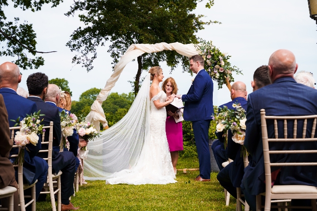 Wedding ceremony outdoors in front of floral moon arch as guests look on. 